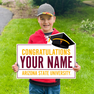 Boy holding lawn sign in yard saying 'Congratulations, Your Name, Arizona State University' with a black graduation cap in the top left corner